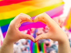 Lesbian making a heart shape with her hands with the LGBTIQ rainbow flag in the background
